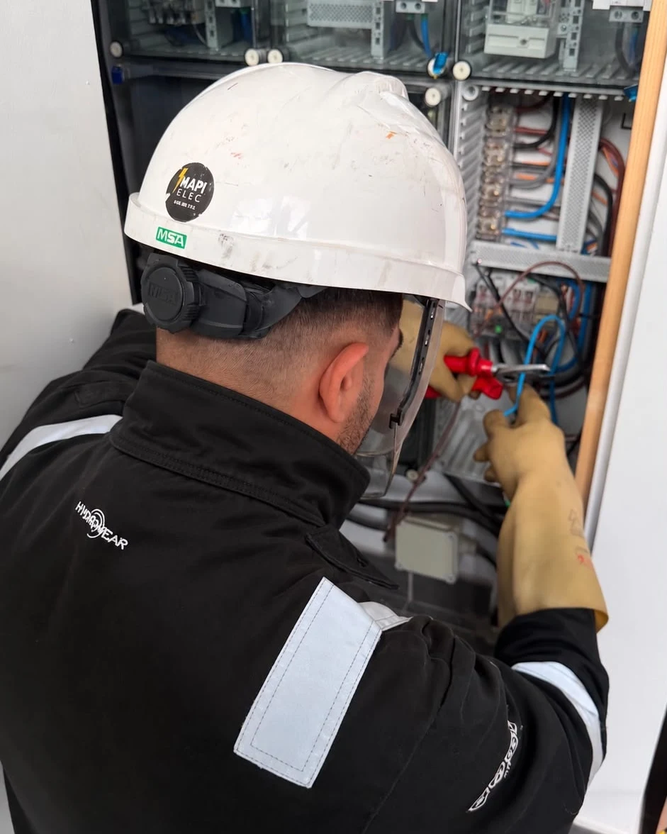 Technicien MAPI Elec en casque, visière et gants isolants, au travail dans une armoire de comptage